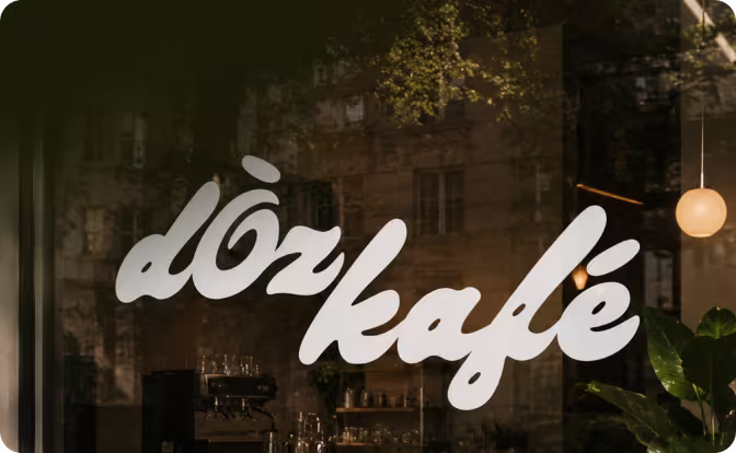 White cursive lettering spelling ’döz kafé’ on a glass window with reflections of buildings and plants.